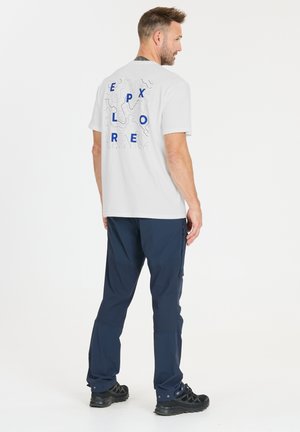 Mann trägt weißes T-Shirt mit blauem "EXPLORE"-Text und Konturlinien, marineblaue Hose und schwarze Wanderschuhe, steht und schaut seitlich.