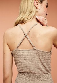 Haut smocké texturé couleur brun clair avec des bretelles croisées et ajustables. La partie arrière présente une coupe ajustée et une bande élastiquée à la taille.