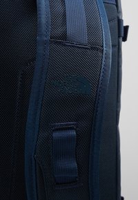 Gros plan d'une sangle de sac à dos noir texturé avec un bord bleu et une boucle cousue, affichant le logo The North Face.