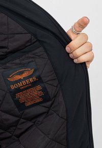 Zwarte bomberjack met een gewatteerde textuur, voorzien van een oranje label met het merk "BOMBERS" en verzorgingsinstructies aan de binnenkant.