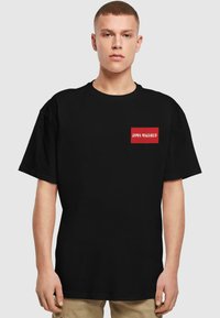 Zwarte katoenen T-shirt met een relaxte pasvorm, voorzien van een rode rechthoekige patch met de tekst "DIMA MAGHREB" in het wit, korte mouwen.