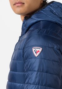 Giacca puffer blu navy con un design trapuntato, caratterizzata da un distintivo al logo Rossignol prominente sulla spalla sinistra e un colletto alto.