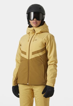 Skijacke in Gelb und Braun mit einem Reißverschluss vorne, Kapuze und dreieckigem Farbblockdesign. Kombiniert mit schwarzen Handschuhen und Helm.
