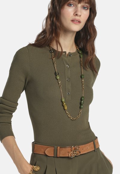 Femme portant un haut à manches longues côtelé vert olive, un long collier de perles dorées, une ceinture marron avec boucle dorée, et un pantalon assorti vert olive.