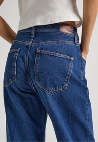 Vaqueros de mezclilla en azul oscuro con un corte relajado, que cuentan con dos bolsillos traseros y un parche de cuero marrón en la cintura. Detalles de costura visibles.