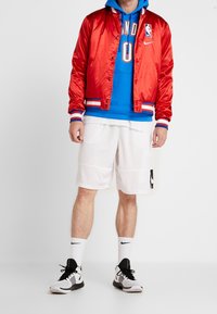 Roter Satin-Bomberjacke mit kontrastierendem blauen Kapuzenpulli, weißen Sportshorts und schwarz-weißen Sneakers. Mit NBA-Logo auf der Jacke.
