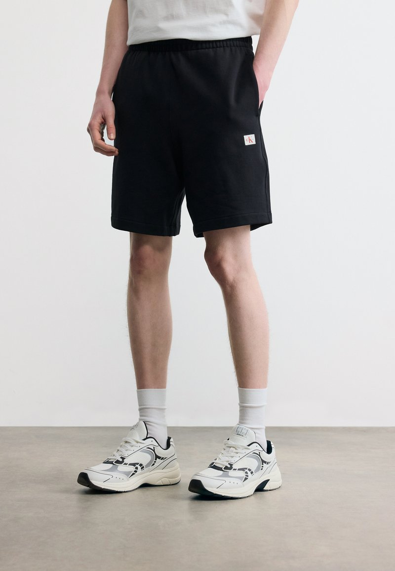 Άτομο που φοράει μαύρα casual σορτς με ένα μικρό λογότυπο στον μηρό, λευκές κάλτσες και λευκά αθλητικά παπούτσια, στέκεται σε λεία επιφάνεια.