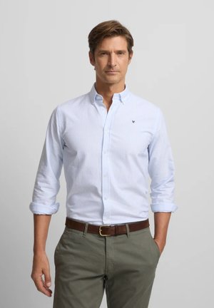 Hombre con camisa azul clara abotonada con las mangas remangadas, pantalones verde oliva y cinturón marrón, de pie con una mano en el bolsillo.