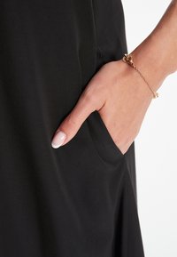 Zwart jurkstof met een gladde textuur en subtiele glans. Een hand met een manicured roze nagel rust in een zijzak, verfraaid met een delicaat armbandje.