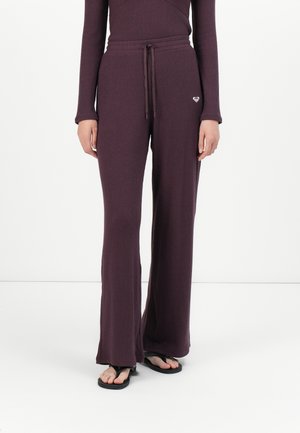 Donna che indossa pantaloni a gamba larga a coste di colore viola scuro con vita regolabile e piccolo emblema a forma di cuore sulla parte anteriore, abbinati a infradito nere.