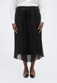 Μαύρη πλισέ μίντι φούστα, συνδυασμένη με λευκό πουκάμισο μακρυμάνικο και μαύρα παπούτσια τύπου slingback με πατεντέ αδιαφανή λεπτομέρεια, σε όρθια στάση μπροστά σε απλό λευκό φόντο.