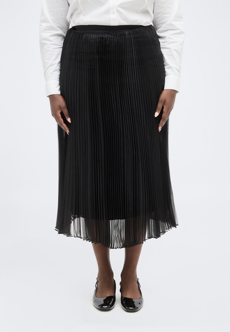 Μαύρη πλισέ μίντι φούστα, συνδυασμένη με λευκό πουκάμισο μακρυμάνικο και μαύρα παπούτσια τύπου slingback με πατεντέ αδιαφανή λεπτομέρεια, σε όρθια στάση μπροστά σε απλό λευκό φόντο.