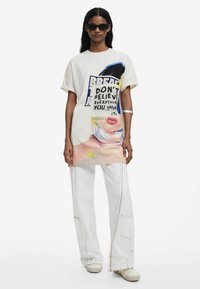 Valkoinen ylisize-t-paita, jossa on graafinen printti, joka sisältää tekstiä ja kasvojen kuvituksen. Yhdistetty laajoihin valkoisiin cargo-housuihin ja vaaleanvärisiin kenkiin.