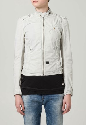 Ljusgrå jacka med ståkrage, framdragkedja och två fickor. Har knappdetaljer och en figursydd design. Bärs över en svart topp.