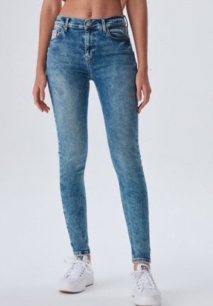 Niebieskie jeansy typu slim fit noszone z białymi tenisówkami na stojącej osobie na jednolitym tle.