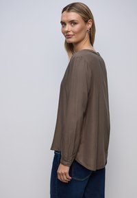 Langærmet, brun bluse lavet af glat stof, med en rund halsudskæring og en let skræddersyet pasform. Matcher med mørkeblå jeans.