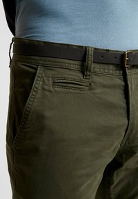 Olivově zelené bavlněné kalhoty s předními kapsami, prošitými detaily a černým koženým opaskem. Hladká textura a přiléhavý design.