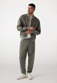 Olijfgroene bomberjack met geribde manchetten, gecombineerd met bijpassende loose-fit broek. Witte sneakers maken de outfit af en bevorderen de casual uitstraling.