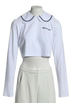 Hvid croppet langærmet top med bølget Peter Pan-krave trimmed med sort og "miu miu" logo på brystet, kombineret med højtaljede hvide bukser.