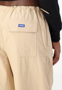 Beige shorts with an elastic waistband, featuring a back pocket with a blue "HUGO" label. Material appears lightweight and textured.