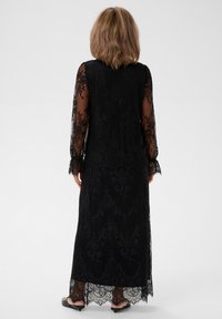 Czarna koronkowa długa sukienka z przezroczystymi długimi rękawami, ozdobiona kwiatowymi wzorami i falbanką u dołu. Posiada nieprzezroczystą podszewkę pod koronką.