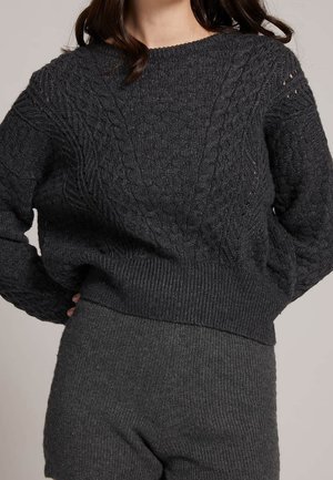 Kvinde iført mørkegrå kabelstrikket sweater med ribbet kant og matchende ribbestrikkede shorts, hænderne på hofterne, neutral baggrund.