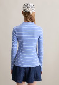 Eine Frau, die mit dem Rücken zu sehen ist, trägt ein hellblaues Langarmoberteil mit weißen horizontalen Streifen, einen dunkelblauen Jeansrock und ein weißes gemustertes Kopftuch.