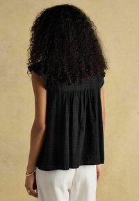 Mustar, lühikeste varrukatega pluus kogutud seljaga, valmistatud kergest kangast. Kombineeritud heledate, lõdvalt istuvate pükstega.