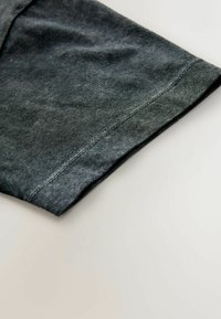 Tejido de mezclilla gris oscuro que muestra un dobladillo doblado con costura verde. La textura es suave con un ligero brillo y detalles de desgaste sutiles.