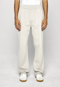 Světlé béžové tepláky s volným střihem, elastickým pasem, bočními kapsami a měkkou texturou; v kombinaci s bílými teniskami.