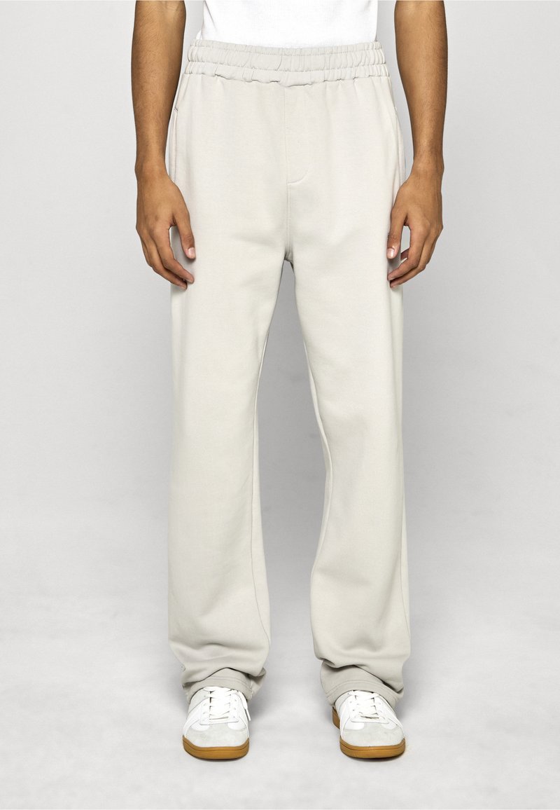 Světlé béžové tepláky s volným střihem, elastickým pasem, bočními kapsami a měkkou texturou; v kombinaci s bílými teniskami.