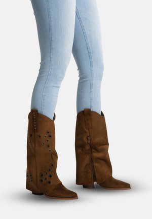 Person iført lyseblå tætsiddende jeans og brune ruskinds cowboystøvler til midt på læggen med dekorative øjne og lynlåse i siden.