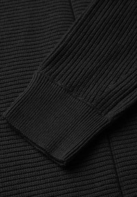 Schwarzer Strickpullover mit gerippter Textur an Ärmeln und Körper, der ein enges Bündchen-Design mit sauber verarbeiteten Kanten zeigt.