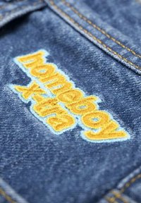 Denim tkanina z modrim odtenkom, ki vsebuje rumeno vezeno besedilo "homeboy". Vidno šivanje v kontrastni barvi.