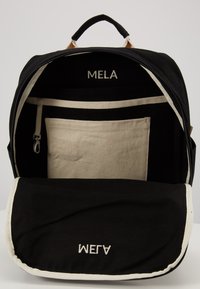 Black canvas backpack with a cream interior, zipper pocket, and "MELA" and "MELA" labels inside. Features a top handle and fold-over flap.