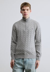 Γκρι πουλόβερ με πλέξη cable, ψηλό γιακά και κλείσιμο με μισό φερμουάρ. Ριμπ μανσέτες και τελείωμα; διαθέτει μικρό λογότυπο στο στήθος.