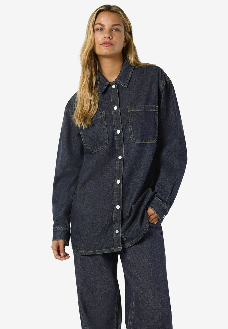 Temno jeans srajca s klasičnim ovratnikom, dvema žepoma na prsih in vidnim šivom. Ima sproščen kroj in zapenjanje na gumbe.