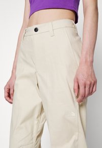 Personne portant un pantalon beige The North Face et un haut court violet, montrant la taille et le haut des jambes sur un fond uni.