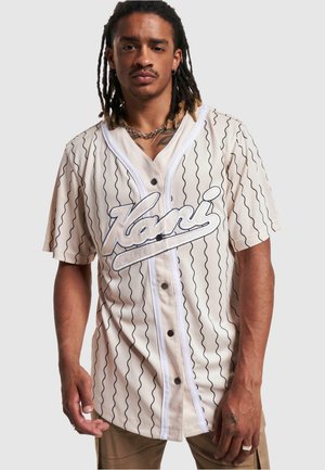 Beige short-sleeve jersey with black wavy stripes and a prominent white embroidered logo. Features button closure and contrast trim.