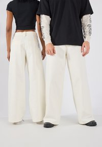 Pantalons larges de couleur crème avec une texture lisse, dotés de deux poches arrière et d'un détail de logo sur le côté gauche près de la taille.