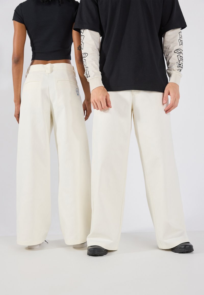 Pantalons larges de couleur crème avec une texture lisse, dotés de deux poches arrière et d'un détail de logo sur le côté gauche près de la taille.