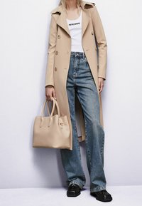 Abrigo trench beige, camiseta blanca sin mangas con texto, vaqueros azules de talle alto, zapatos negros y un bolso de cuero beige suave.