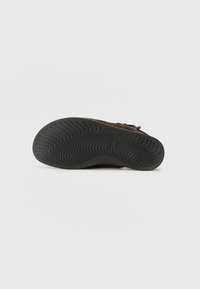 Semelle d'une chaussure marron avec une semelle en caoutchouc noir texturée présentant des motifs ondulés pour l'adhérence, montrée sur un fond gris uni.