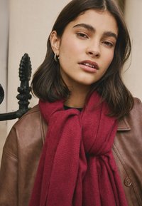 Weinroter Schal aus weichem Stoff, locker vorne geknotet, getragen über einer braunen Lederjacke mit sichtbaren strukturierten Details.