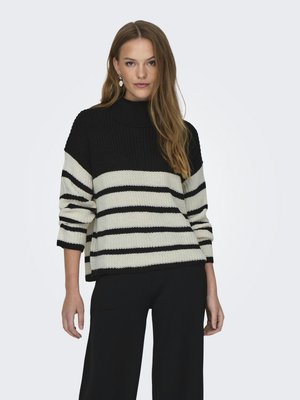 Pull en maille noir et crème à rayures, avec un col montant et des manches larges, surface texturée, porté avec un pantalon large noir.