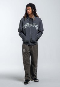 Gray zippered hoodie with large white graphic text. Loose olive green pants featuring embroidered accents. Black shoes with white soles.