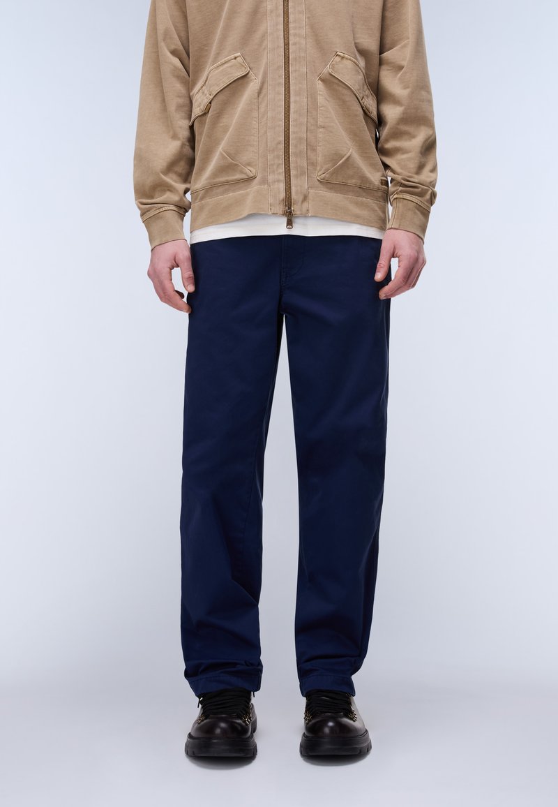 Hombre con pantalones azul marino de pierna recta, zapatos negros y una chaqueta con cremallera color caqui con bolsillos con solapa, posando frente a un fondo gris liso.