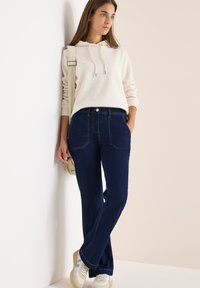 Jeans in denim blu scuro con vestibilità rilassata, dotati di due tasche anteriori e cuciture a contrasto. Abbinati a una felpa leggera color beige.