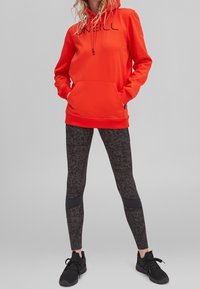 Personne portant un sweat à capuche orange vif avec poche avant, des leggings noirs à motifs, et des chaussures de sport noires sur un fond gris.