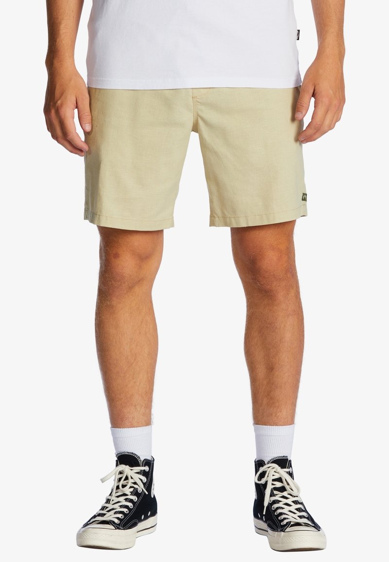 Shorts beige clair en tissu texturé, coupe droite, avec un petit patch logo. Associés à des baskets montantes noires et des chaussettes blanches mi-mollet.